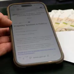 Dłoń trzyma smartfon z informacją o Krajowym Systemie e-Faktur. Obok leżą banknoty, sugerując zwrot VAT.