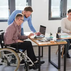 Dwóch mężczyzn pracuje przy biurku. Jeden siedzi na wózku inwalidzkim, drugi stoi obok. Trzeci mężczyzna pracuje przy laptopie przy innym biurku. To przykład efektywnego czasu pracy osoby niepełnosprawnej.