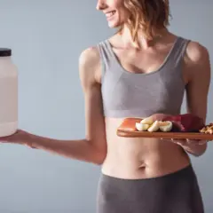 Femme souriante tenant un pot de protéines et une assiette avec viande, œufs, fromage et noix. Idéal pour le calcul protéine par jour femme.