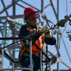 Elektryk w kasku i kamizelce odblaskowej pracuje na wysokości przy słupie energetycznym.