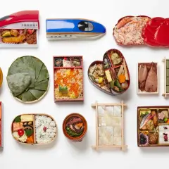 A variety of Japanese bento boxes are displayed, including train-shaped ones, a heart-shaped bento, and those filled with seafood, rice, and other delicacies.
