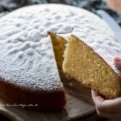 Una fetta di torta margherita soffice, cosparsa di zucchero a velo, viene sollevata da una mano.