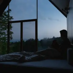 Mann sitzt im Bett, blickt aus dem Fenster in die Dämmerung. Ein ruhiger Moment, der den schlaf wach rhythmus widerspiegelt.