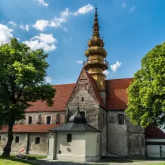 Kamienna świątynia z żółtą wieżą i czerwonym dachem, otoczona zielenią. W oddali widać fragmenty zabudowań, przypominające krajobraz wokół **zamku Krzyżtopór**.