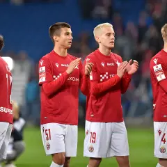 Piłkarze w czerwonych koszulkach i białych spodenkach klaszczą na boisku.