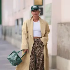 Modna kobieta w beżowym płaszczu, białym topie i spodniach w panterkę. Zielona torebka i czapka dopełniają stylizację.
