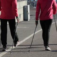 Dwie osoby w czerwonych kurtkach i czarnych spodniach idą z kijkami do nordic walking. Pokazują, jak ustawić kijki do chodzenia.