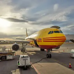 Żółty samolot DHL z czerwonymi pasami na płycie lotniska, gotowy do lotu do USA.
