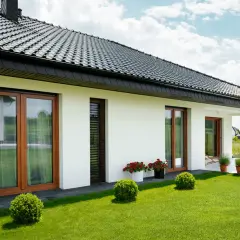 Nowoczesny dom z białymi ścianami, drewnianymi oknami i czarnym dachem. Elewacja w jasnym kolorze, idealnie komponuje się z zielonym ogrodem, prezentując najmodniejsze kolory elewacji.