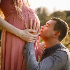 Ciepła sesja ciążowa z mężem. On klęczy i całuje brzuch ukochanej, która delikatnie go obejmuje.