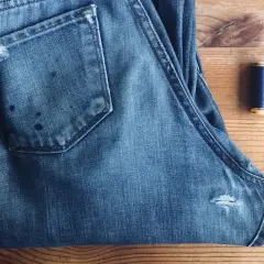 A pair of blue jeans with a small rip on the back pocket and a spool of navy thread, ready for how to patch jeans.