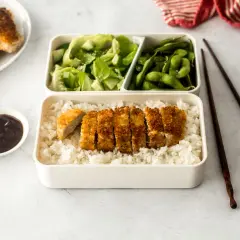 A delicious pork katsu bento box with rice, salad, edamame, and sauce.