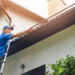 Man on ladder cleaning gutters. This task should be done at least twice a year to prevent clogs.