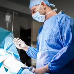 Chirurg w niebieskim fartuchu operacyjnym i masce trzyma narzędzia podczas zabiegu.