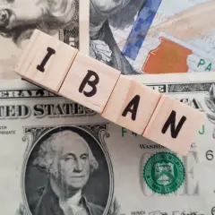 Drewniane klocki z napisem IBAN leżą na banknotach dolarowych.