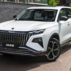 Biały, chiński SUV z imponującym grillem i napisem "HUGE" na masce, zaparkowany na podjeździe z kostki brukowej.