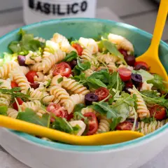 Ensalada de pasta espectacular con fusilli, tomates cherry, aceitunas negras y rúcula, servida en un cuenco azul con cucharas amarillas.