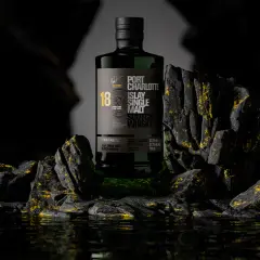 Bouteille de whisky Port Charlotte 18 ans d'âge, produit par la distillerie Bruichladdich, posée sur des rochers sombres aux reflets dorés.