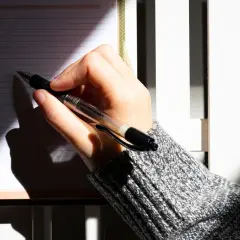 Dłoń pisze w notatniku, planując letnie podróże. Cień pada na kartki, jakby to były święta w lipcu.