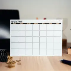 Pusta plansza kalendarza z niemieckimi dniami tygodnia na biurku, obok laptopa i rośliny.