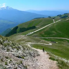 A person walks on a gravel path on a green mountain slope with a road winding through the hills and a snow-capped mountain in the distance.