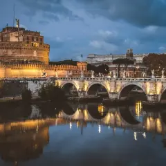 Le Castel Sant'Angelo, illuminé la nuit, se reflète dans le Tibre, avec un pont arqué en avant-plan.