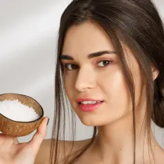 Mujer sonríe sosteniendo una cáscara de coco con gránulos blancos, ideal para el aceite de coco para el pelo.