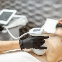 Pacjent leży na stole, a specjalista w czarnej rękawiczce trzyma urządzenie do liposukcji ultradźwiękowej na jego brzuchu.