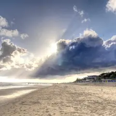 Plaża na wyspie w Niemczech. Słońce przebija się przez burzowe chmury, oświetlając piaszczysty brzeg i rzędy kolorowych budek plażowych.