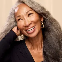 Mujer sonríe con cabello largo y ondulado, mostrando una hermosa transición a canas.
