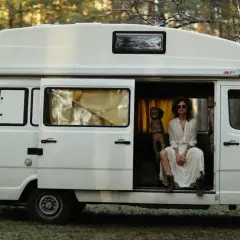 Kobieta w białej sukience siedzi w otwartych drzwiach białego kampera, otoczona lasem. Idealne polskie caravaning miejscówki na wakacje.