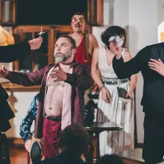 Aktorzy na scenie, w tym mężczyzna z brodą w rozpiętej koszuli, prezentują swój repertuar. Widownia w napięciu obserwuje rozwój wydarzeń.