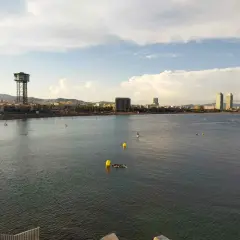 Vista de la playa y el mar con el icónico hotel vela al fondo. Personas disfrutan del agua en tablas de paddle surf.