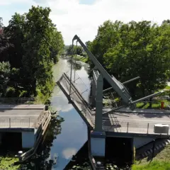 Podniesiony most zwodzony w Nowym Dworze Gdańskim, nad kanałem otoczonym zielenią.