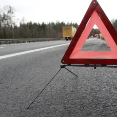 Czerwony trójkąt ostrzegawczy stoi na poboczu autostrady, sygnalizując niebezpieczeństwo. W tle widać ciężarówkę i inne pojazdy.