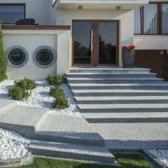 Nowoczesne schody zewnętrzne z betonu dekoracyjnego, otoczone białym żwirem i zielenią, prowadzą do eleganckich drzwi wejściowych.