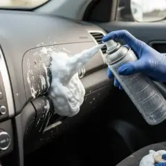 Mano con guanto blu spruzza schiuma da barba per pulire sedili auto e cruscotto.
