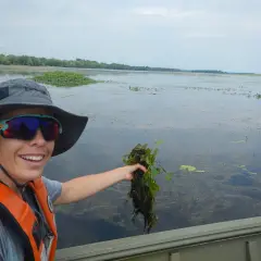 Une femme en gilet de sauvetage tient une plante aquatique, peut-être une châtaigne d'eau, dans un lac.