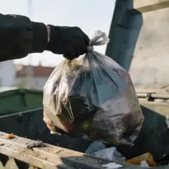 Ręka w czarnej rękawiczce wrzuca worek ze śmieciami do kontenera. Zastanawiamy się, ile zapłacimy za wywóz śmieci.