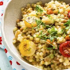Un plato de pasta pequeña, que es semola, con tomates cherry asados y rodajas de limón, adornado con hierbas frescas.