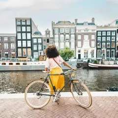 Kobieta z rowerem na tle amsterdamskich kamienic i kanału. Idealne miejsce na odkrywanie tras rowerowych Europa.