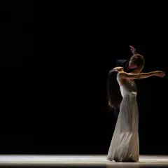 Zwei Tänzer des Tanztheater Wuppertal in inniger Umarmung auf dunkler Bühne. Die Frau im weißen Kleid neigt den Kopf zurück, der Mann beugt sich über sie.