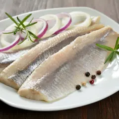 Trzy filety śledzi w occie, udekorowane krążkami cebuli, gałązkami rozmarynu i kolorowym pieprzem, na białym talerzu.