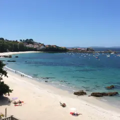 La **playa de Aguete** luce un día soleado con aguas cristalinas y barcos fondeados. Gente disfruta de la arena blanca y el mar azul.