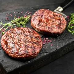 Dwa soczyste, grillowane kotlety na burgery na drewnianej desce z ziołami i różowym pieprzem.