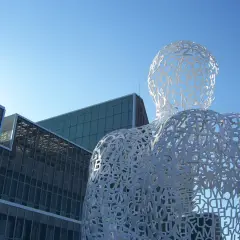 Biała, ażurowa rzeźba postaci z liter, przypominająca monumentalizm, na tle nowoczesnych budynków i błękitnego nieba.