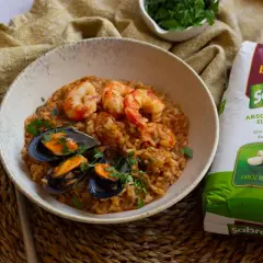 Un plato de arroz con gambas congeladas y mejillones, decorado con perejil fresco. Al lado, un paquete de arroz redondo Sabroz.