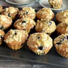 Pyszne babeczki z jagodami, posypane kruszonką, ułożone na ciemnej tacy. Idealne na śniadanie lub deser.