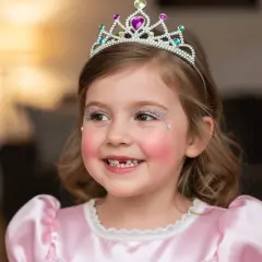A young girl in a pink princess dress and tiara smiles, with glitter makeup on her eyes and cheeks.