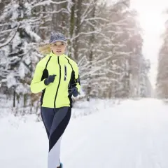 Kobieta biegnie przez zaśnieżony las, mimo że jest rzs a zimno. W neonowej kurtce i czapce stawia czoła mrozowi.
